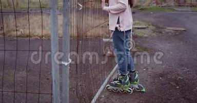 穿着溜冰鞋的年轻女孩用手拿着生锈的栅栏，透过它们看。 女孩穿着粉色背心